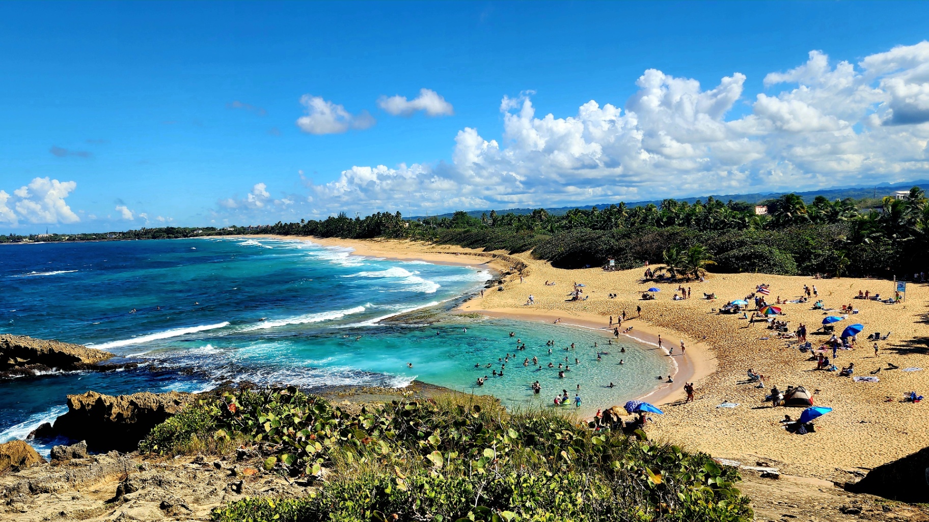 Aerial view of a golden sand beach on Puerto Rico's north coast with turquoise water, beachgoers, and lush green coastline under a bright blue sky